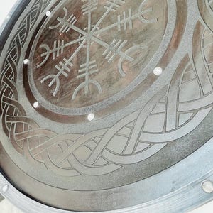 May include: A circular, metallic shield with intricate, engraved Viking-inspired designs. The shield features a central symbol surrounded by knotwork and a border with rivets. The color is a muted silver-gray, with a slightly weathered appearance.