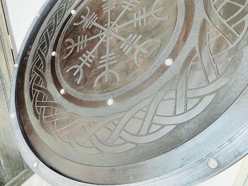May include: A circular, metallic shield with intricate, engraved Viking-inspired designs. The shield features a central symbol surrounded by knotwork and a border with rivets. The color is a muted silver-gray, with a slightly weathered appearance.