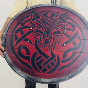 May include: Round wooden shield with a red background and a black wolf design. The shield has a metal rim and is held by a person. The wolf design is intricate and detailed, with a focus on the animal's face.