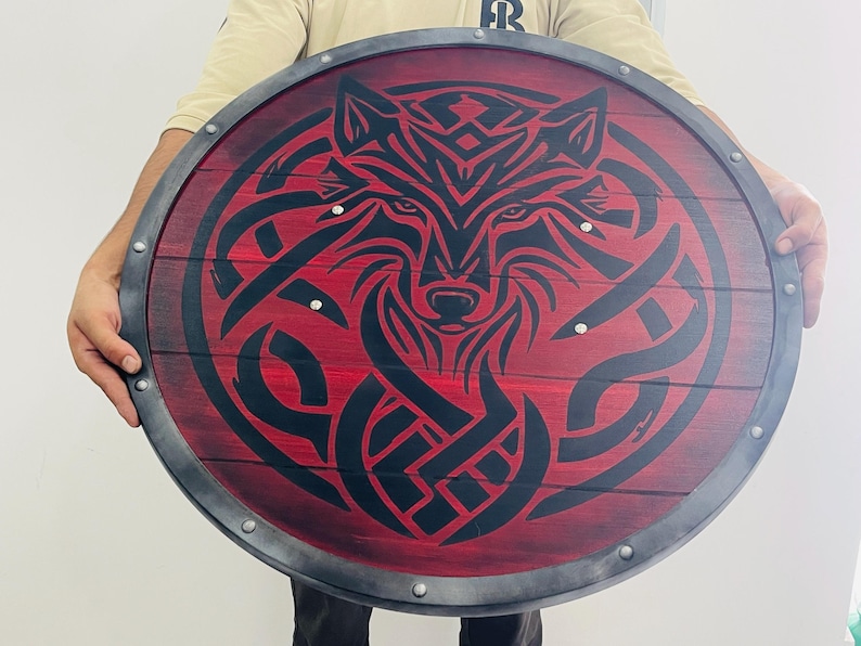 May include: Round wooden shield with a red background and a black wolf design. The shield has a metal rim and is held by a person. The wolf design is intricate and detailed, with a focus on the animal's face.