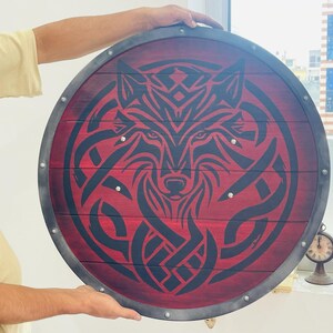 May include: Round wooden shield with a red background and a black wolf design. The shield has a metal rim and rivets. The wolf design is a stylized, symmetrical illustration. The shield is held by a person.