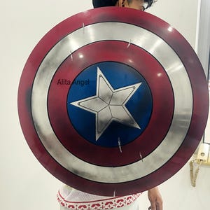 May include: A round shield with a red, white, and blue color scheme, featuring a white star in the center. The shield has a distressed, weathered appearance. The text "Alita Angel" is visible on the shield. This item is a prop or collectible.