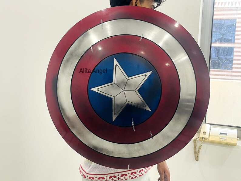 May include: A round shield with a red, white, and blue color scheme, featuring a white star in the center. The shield has a distressed, weathered appearance. The text "Alita Angel" is visible on the shield. This item is a prop or collectible.