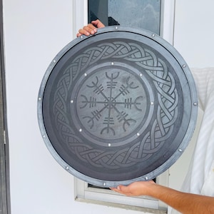 May include: A round, gray shield with a detailed Viking design. The shield features a central symbol surrounded by intricate knotwork patterns. Silver rivets secure the outer rim. A decorative piece for historical enthusiasts.