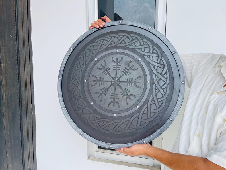 May include: A round, gray shield with a detailed Viking design. The shield features a central symbol surrounded by intricate knotwork patterns. Silver rivets secure the outer rim. A decorative piece for historical enthusiasts.