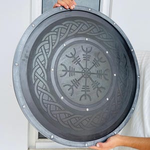 May include: A round, gray shield with a dark gray center featuring a Viking design. The shield has a silver-colored trim and is held by a person. The shield's center has a circular pattern with a symbol in the middle.
