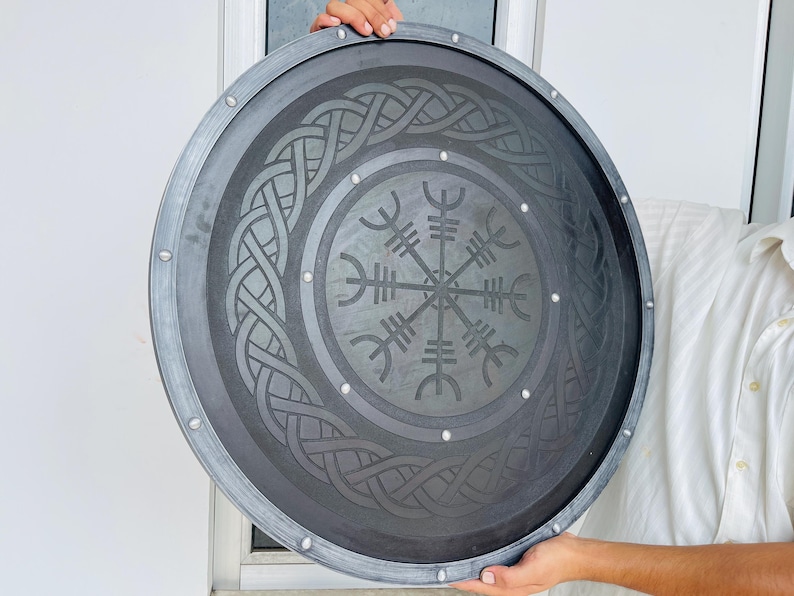 May include: A round, gray shield with a dark gray center featuring a Viking design. The shield has a silver-colored trim and is held by a person. The shield's center has a circular pattern with a symbol in the middle.