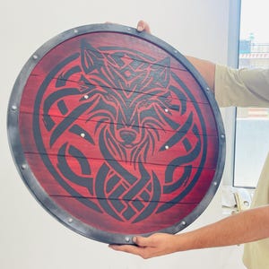 May include: Round wooden shield with a red background and a black wolf design. The shield has a dark gray metal rim and is held by a person. The wolf design is in a Celtic knot style.