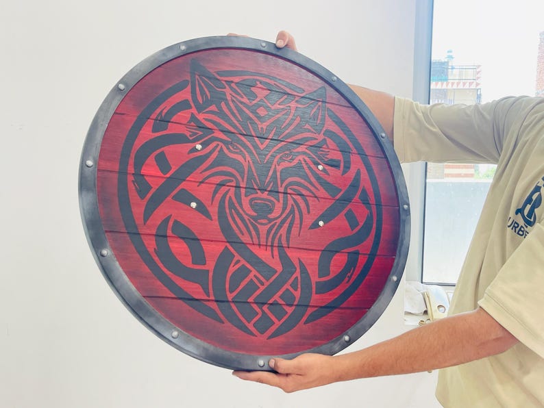 May include: Round wooden shield with a red background and a black wolf design. The shield has a dark gray metal rim and is held by a person. The wolf design is in a Celtic knot style.
