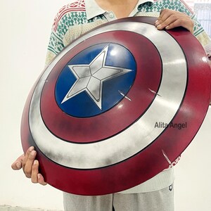 May include: A round shield with a red, white, and blue color scheme, featuring a white star in a blue circle at the center. The shield has a metallic silver outer ring and visible scratches. The shield is held by a person.