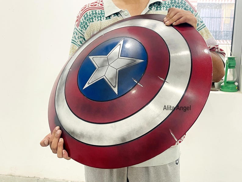 May include: A round shield with a red, white, and blue color scheme, featuring a white star in a blue circle at the center. The shield has a metallic silver outer ring and visible scratches. The shield is held by a person.