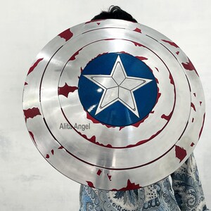 May include: A silver-colored shield with a blue central circle featuring a white star. The shield has red accents around the edge, with a distressed, battle-worn appearance. The shield is round and appears to be a prop or costume accessory.