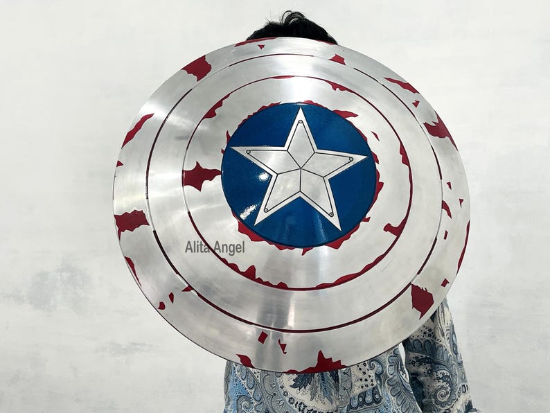 May include: A silver-colored shield with a blue central circle featuring a white star. The shield has red accents around the edge, with a distressed, battle-worn appearance. The shield is round and appears to be a prop or costume accessory.