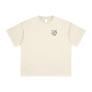 Puede incluir: Camiseta de color crema con un pequeño gráfico en blanco y negro de un personaje de dibujos animados en la parte superior izquierda del pecho. La camiseta tiene mangas cortas y cuello redondo. La tela parece suave y cómoda.