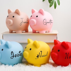 Könnte beinhalten: Fünf bunte Sparschweine in Rosa, Blau, Gelb und Rot. Auf jedem Schwein steht ein Name in Schreibschrift: Javek, Garth, Jodie, Hazel und Novia. Sie stehen auf einer weißen Oberfläche, ideal für Kinder.