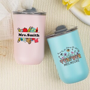 May include: Two insulated tumblers, one pink and one blue, with black lids. The pink tumbler has a teacher-themed graphic with the name "Mrs. Smith." The blue tumbler says "TEACHER MRS. MOORE" with a floral design.