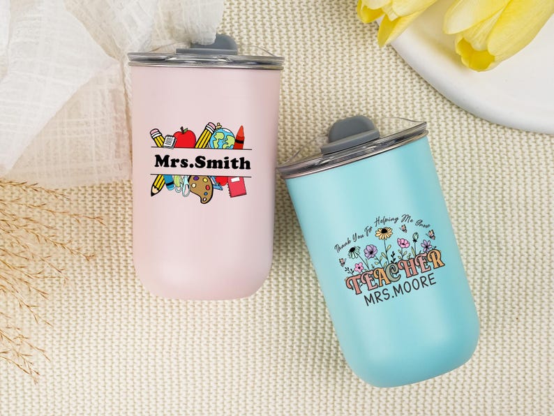 May include: Two insulated tumblers, one pink and one blue, with black lids. The pink tumbler has a teacher-themed graphic with the name "Mrs. Smith." The blue tumbler says "TEACHER MRS. MOORE" with a floral design.