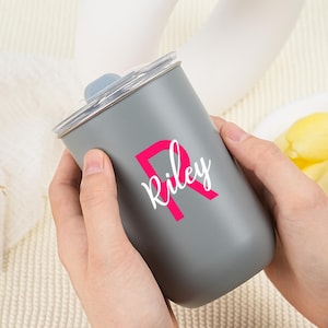 May include: A gray tumbler with a clear lid is held by two hands. The tumbler has a pink letter "R" with the name "Riley" in white script. The tumbler is a cylindrical shape.