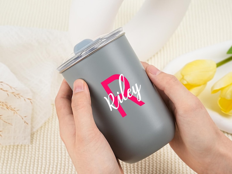 May include: A gray tumbler with a clear lid is held by two hands. The tumbler has a pink letter "R" with the name "Riley" in white script. The tumbler is a cylindrical shape.