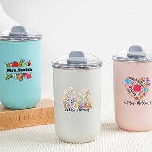 May include: Three insulated tumblers with clear lids and gray tops. The tumblers are light blue, white, and pink. Each tumbler has a different design and the name of a teacher. The light blue tumbler has the name "Mrs. Smith".