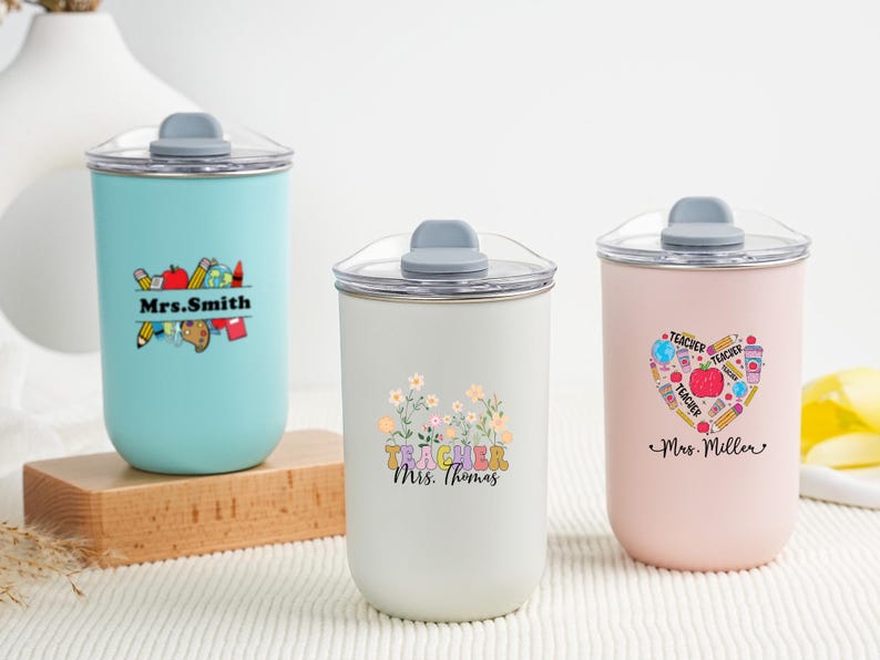 May include: Three insulated tumblers with clear lids and gray tops. The tumblers are light blue, white, and pink. Each tumbler has a different design and the name of a teacher. The light blue tumbler has the name "Mrs. Smith".