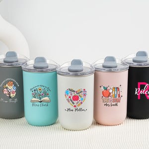 Könnte beinhalten: Fünf isolierte Becher mit transparenten Deckeln in verschiedenen Farben: Grau, Türkis, Weiß, Rosa und Schwarz. Jeder Becher hat ein einzigartiges Design, darunter Blumenmuster, ein Apfel und personalisierter Text. Die Becher sind für Lehrer konzipiert.