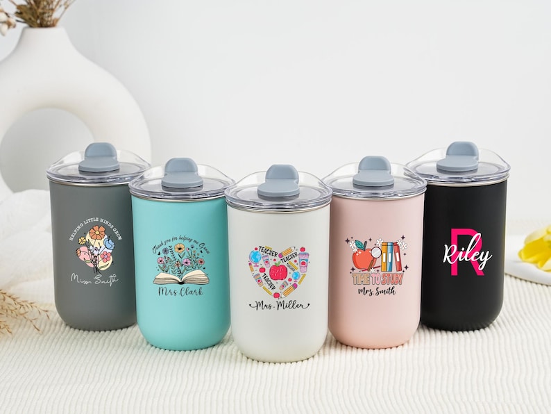 May include: A collection of five insulated tumblers with clear lids. The tumblers are in various colors: gray, teal, white, pink, and black. Each tumbler features a unique design, including floral motifs, an apple, and personalized text. The tumblers are designed for teachers.