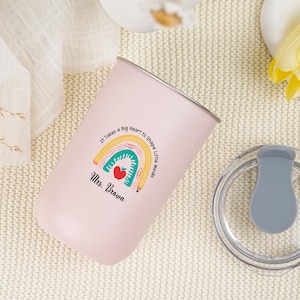 May include: A pink insulated tumbler with a silver rim and a clear lid. The tumbler features a colorful rainbow design with a red apple and the text "It Takes a Big Heart to Shape Little Minds. Mrs. Brown."