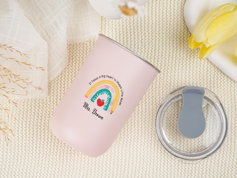 May include: A pink insulated tumbler with a silver rim and a clear lid. The tumbler features a colorful rainbow design with a red apple and the text "It Takes a Big Heart to Shape Little Minds. Mrs. Brown."