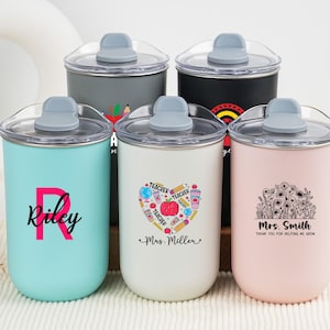 May include: A collection of insulated tumblers in various colors, each with a clear lid and gray handle. One tumbler is light blue with the name "Riley", another is white with a heart design, and a third is pink with a floral design. The tumblers are designed for teachers.