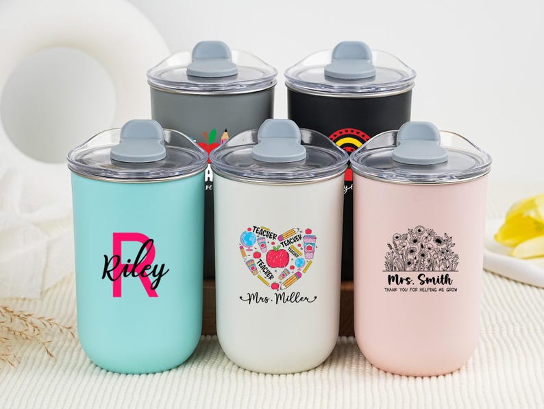 May include: A collection of insulated tumblers in various colors, each with a clear lid and gray handle. One tumbler is light blue with the name "Riley", another is white with a heart design, and a third is pink with a floral design. The tumblers are designed for teachers.