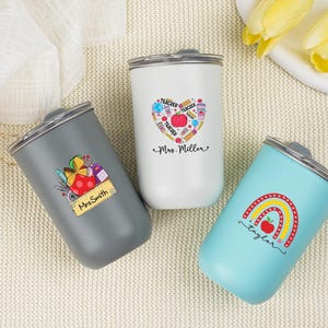 May include: Three insulated tumblers with silver lids. One is gray with a teacher-themed graphic and the name "Mrs. Smith". Another is white with a heart-shaped teacher graphic and the name "Mrs. Miller". The third is light blue with a rainbow graphic and the name "Taylor".