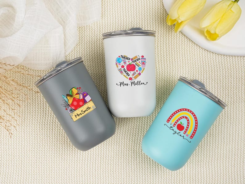 May include: Three insulated tumblers with silver lids. One is gray with a teacher-themed graphic and the name "Mrs. Smith". Another is white with a heart-shaped teacher graphic and the name "Mrs. Miller". The third is light blue with a rainbow graphic and the name "Taylor".
