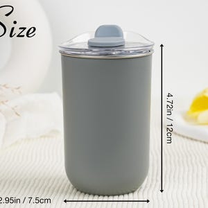 May include: A gray tumbler with a clear lid and a gray flip-top closure. The tumbler is 4.72 inches (12 cm) tall and 2.95 inches (7.5 cm) wide. The word "Size" is written in cursive on the upper left.