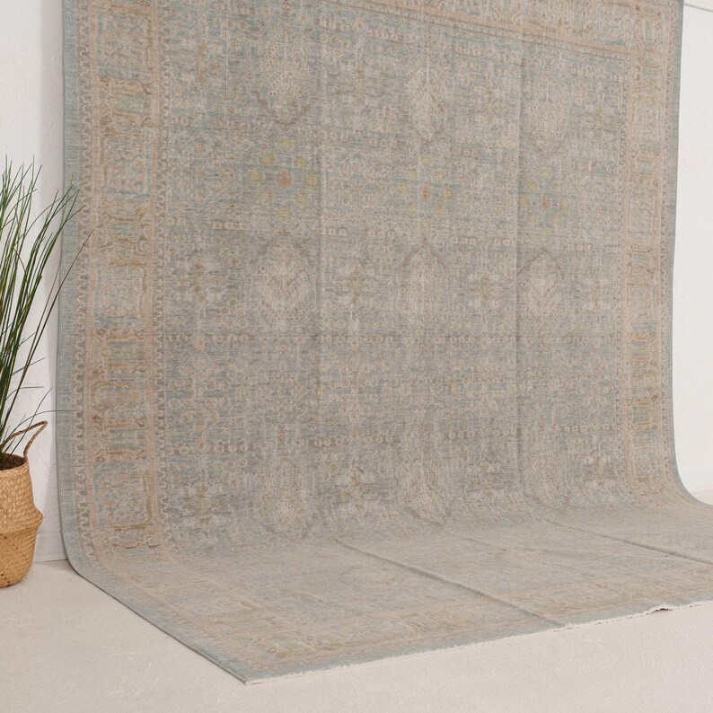 May include: A large area rug featuring a faded, intricate pattern in shades of light blue, beige, and soft gray. The design includes a central medallion and a wide, detailed border with repeating geometric and floral motifs across the surface.