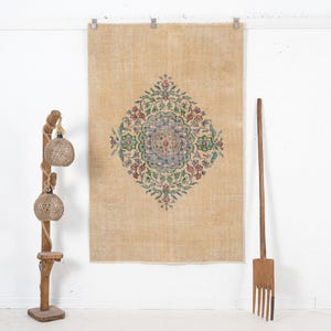 May include: A rectangular area rug in a light tan color featuring a central diamond-shaped medallion with intricate floral and leaf patterns in shades of blue, green, and red. The rug is hung vertically against a plain white wall surface.