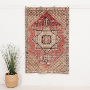 Puede incluir: Una alfombra rectangular con un medallón geométrico central en tonos rojo, beige y gris carbón oscuro. La alfombra presenta un patrón simétrico con motivos florales y un borde decorativo, colgada verticalmente sobre una pared blanca.