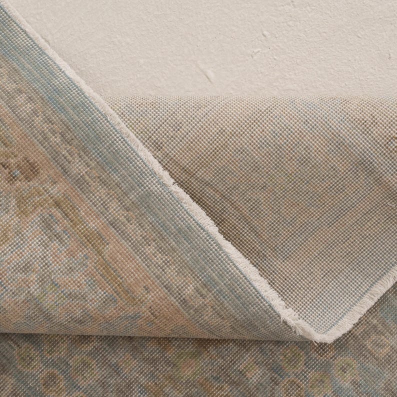 May include: A folded area rug showing a patterned top surface in muted shades of blue, beige, and brown, contrasted with a plain, light beige woven backing. The fabric texture is visible, highlighting the weave of the rug's underside and edges.