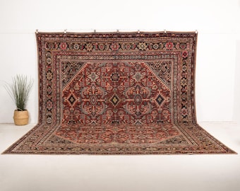 May include: A large rectangular area rug featuring a deep red background with intricate geometric and floral patterns in navy blue, cream, and black. The rug has a wide, detailed border with repeating motifs and a central medallion design.