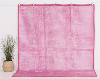 May include: A large, rectangular pink fabric backdrop featuring a mottled, distressed texture throughout the surface. The fabric is finished with a solid pink border along all four edges and is suspended from a horizontal rod by metal clips.