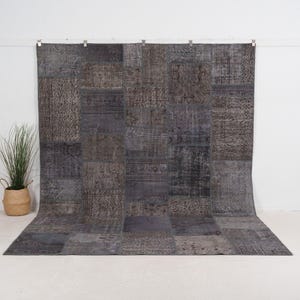 May include: A large, rectangular patchwork rug in shades of gray and brown. The rug is made up of various rectangular and square pieces, each with a unique pattern. The rug is displayed against a white background, with a potted plant in a woven basket to the left.