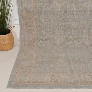 May include: A large area rug featuring a faded, intricate floral and geometric pattern in muted shades of dusty blue, soft beige, and cream. The rug has a wide, detailed border and a central field with a repeating, weathered-look design.