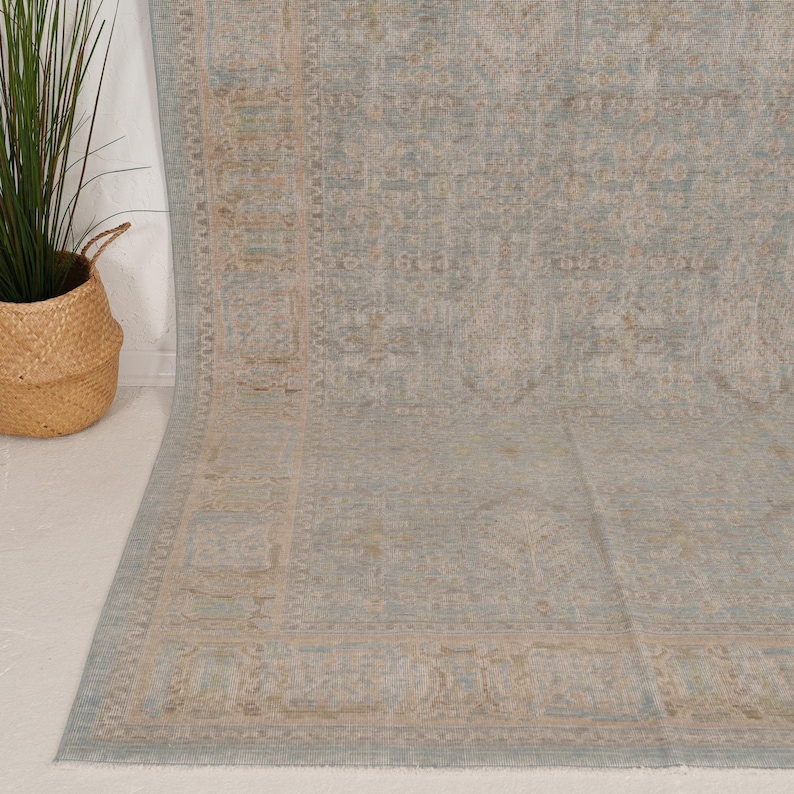 May include: A large area rug featuring a faded, intricate floral and geometric pattern in muted shades of dusty blue, soft beige, and cream. The rug has a wide, detailed border and a central field with a repeating, weathered-look design.