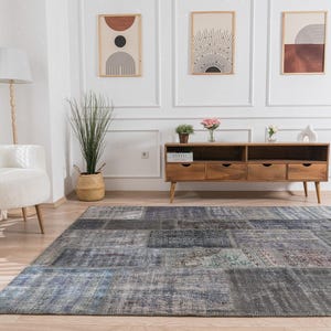 May include: A living room scene featuring a large, rectangular, patchwork-style rug in shades of gray and blue. The room includes a wooden console with drawers, a white armchair, and decorative wall art. A floor lamp and a small side table are also visible.