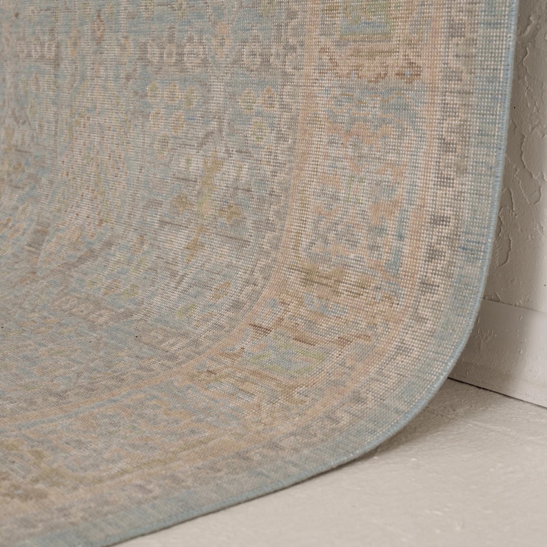 May include: A woven area rug featuring a light blue base color with intricate, faded patterns in shades of beige, tan, and soft gray. The rug has a distinct border design and a textured surface, shown partially curled to reveal its flexible construction.
