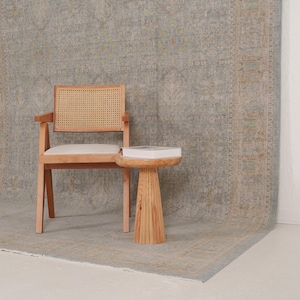 May include: A light wood armchair with a woven cane backrest and a white upholstered seat, positioned next to a small, round, pedestal-style side table made of light wood, which holds a white book, all set against a large, patterned area rug.
