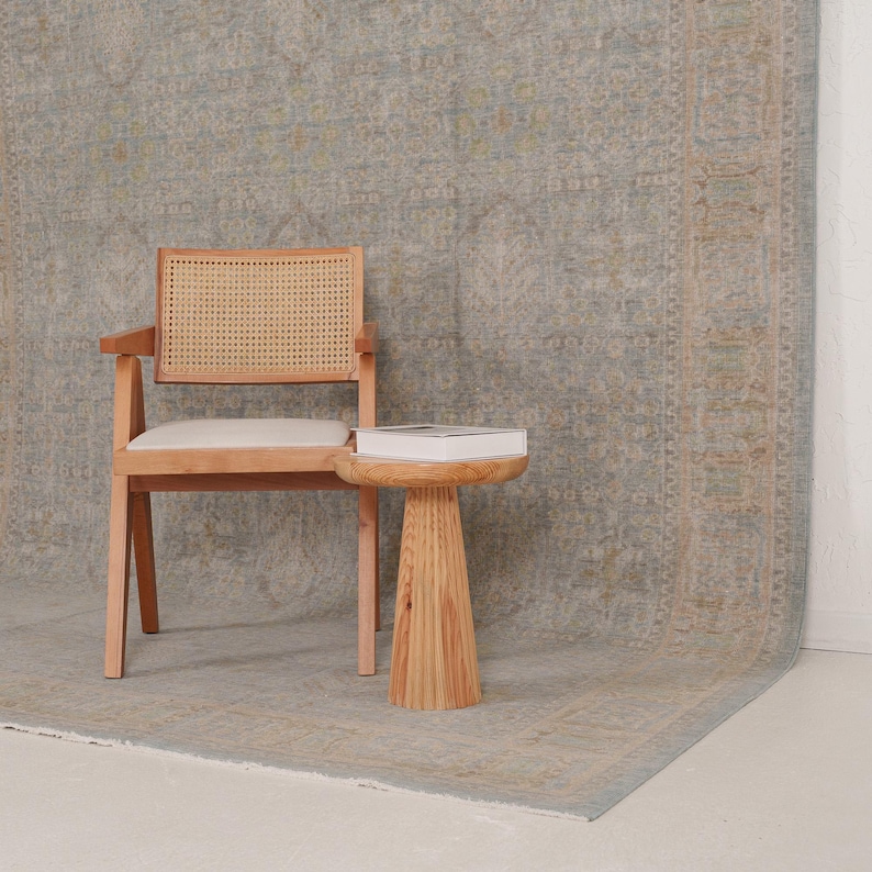 May include: A light wood armchair with a woven cane backrest and a white upholstered seat, positioned next to a small, round, pedestal-style side table made of light wood, which holds a white book, all set against a large, patterned area rug.