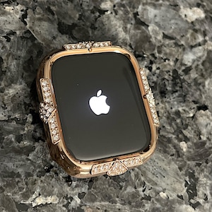 Può includere: Una cassa per Apple Watch in oro rosa con una lunetta incrostata di cristalli simili a diamanti. Il quadrante dell'orologio è nero e mostra il logo Apple.