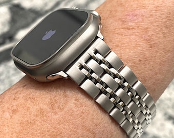 以下が含まれることがあります： ステンレス製のバンドが付いたシルバーのApple Watch。時計の文字盤は黒で、Appleのロゴが表示されています。