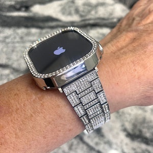 Op de afbeelding: Een zilveren Apple Watch met een band en bezel bezet met diamanten. De wijzerplaat van het horloge is zwart en toont het Apple-logo.